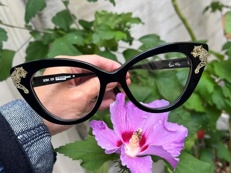 A hand holding a black Francis Klein cat-eye frame with crystals set at the temple